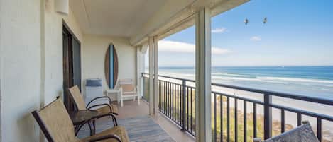 A balcony with a view of the ocean, chairs, and a surfboard leaning against the wall.