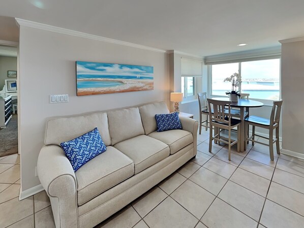 A living room with a beige sofa, two blue patterned pillows, a painting of a beach scene, a dining table with chairs, and a view of the ocean through a large window.