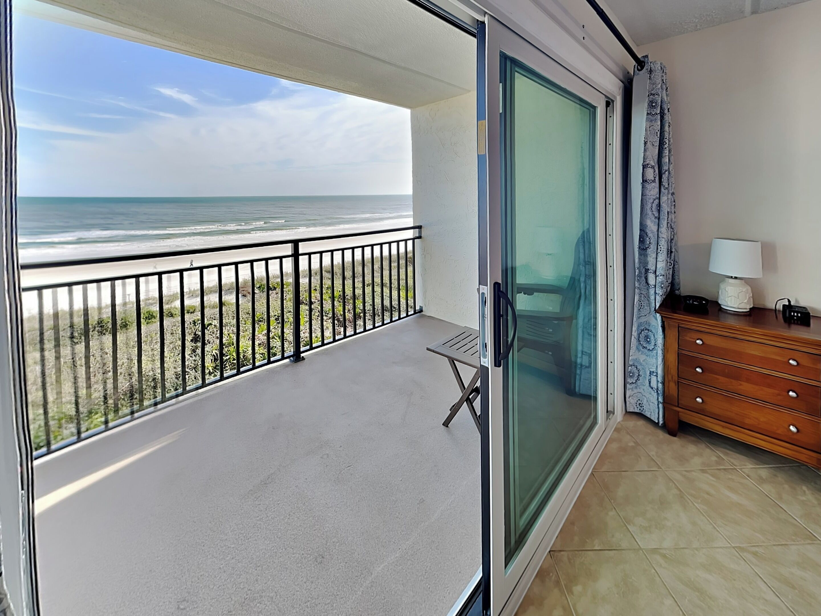 A balcony with a view of the ocean, a sliding glass door, a small table, and a wooden dresser with a lamp.