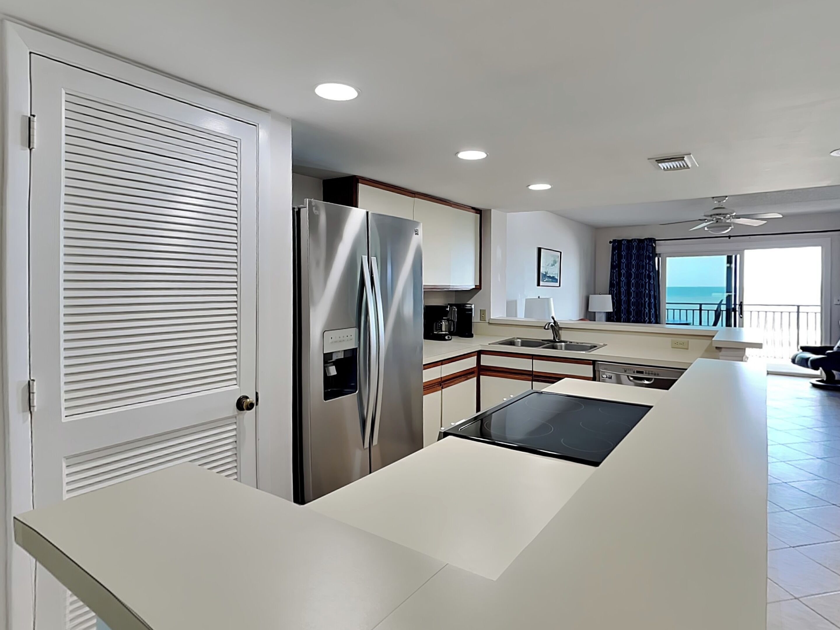 A modern kitchen with a stainless steel refrigerator, a built-in water dispenser, and a black induction cooktop. There is a window with a view of the ocean and a balcony with a view of the sea.