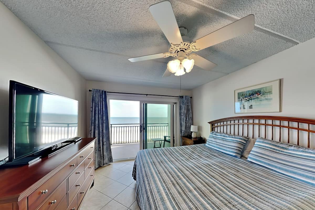 Ein Hotelzimmer mit Bett, Fernseher, Deckenventilator, Balkon mit Meerblick und einem Bild an der Wand.