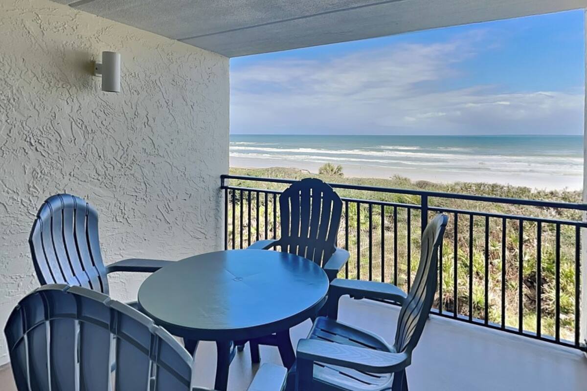 Ein Balkon mit Tisch und Stühlen mit Blick auf einen Strand, Wellen und einen klaren Himmel.