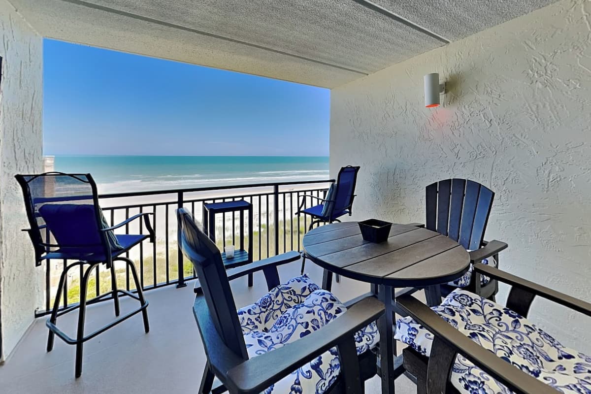 Ein Balkon mit Blick auf den Strand, ausgestattet mit einem Tisch und Stühlen mit gemusterten Kissen.