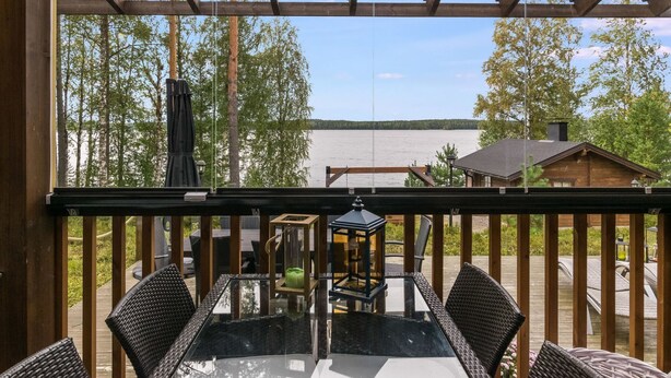 Un terrazzo in legno con tavolo in vetro e sedie in vimini, con vista su un lago e una foresta.