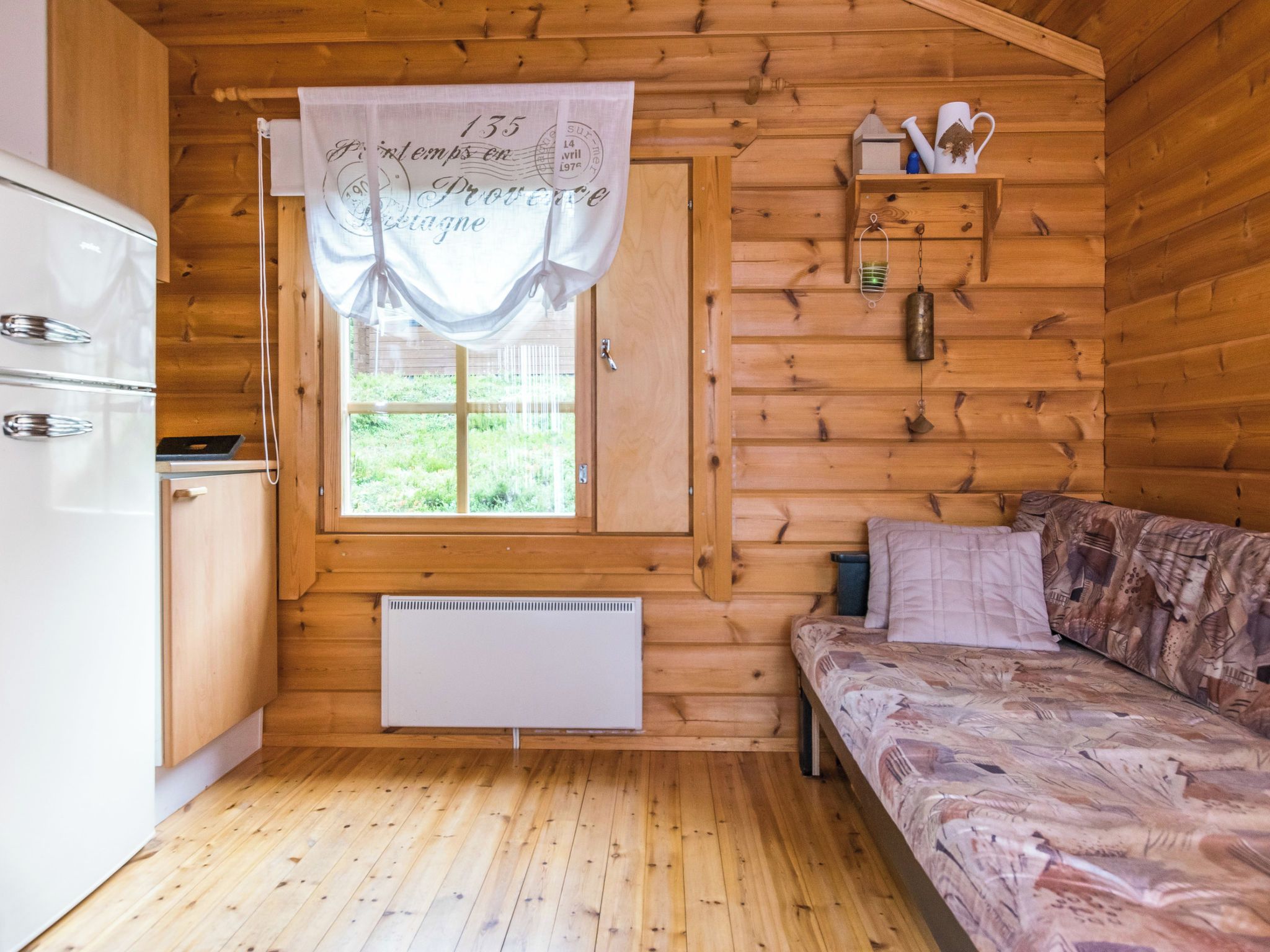 A cozy cabin interior with a bed, a small shelf with decorative items, a window with a curtain, and a white radiator.