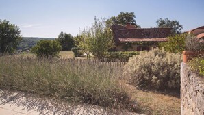 A rustic house with a tiled roof, surrounded by dense shrubbery and a stone wall.