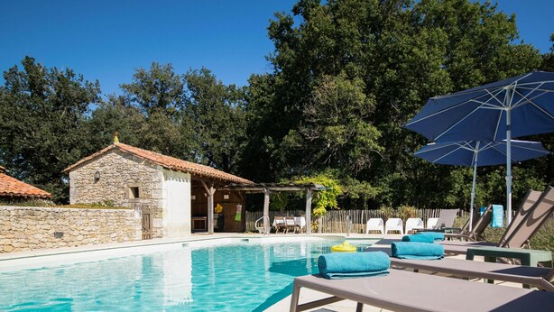 A swimming pool with lounge chairs and an umbrella, surrounded by trees and a stone building.