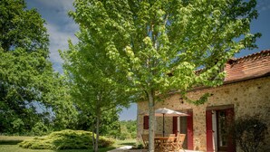 A stone building with a red door, a patio with a table and chairs, and a tree in the foreground. - Authentic gîte on estate with swimming pool (Issac)