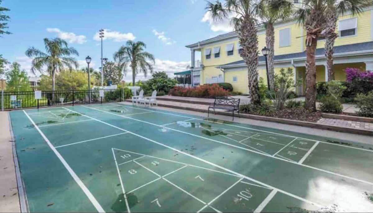 Uma quadra de jogo de shuffleboard com quadrados numerados, cercada por uma grade, palmeiras e um prédio com varandas.