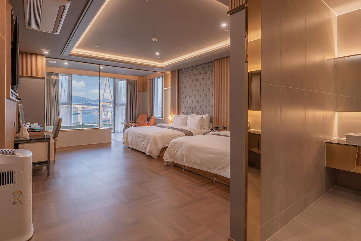 Habitación de hotel moderna con dos camas, un ventanal grande con vista a la ciudad, un escritorio con silla y un baño con lavamanos y espejo.