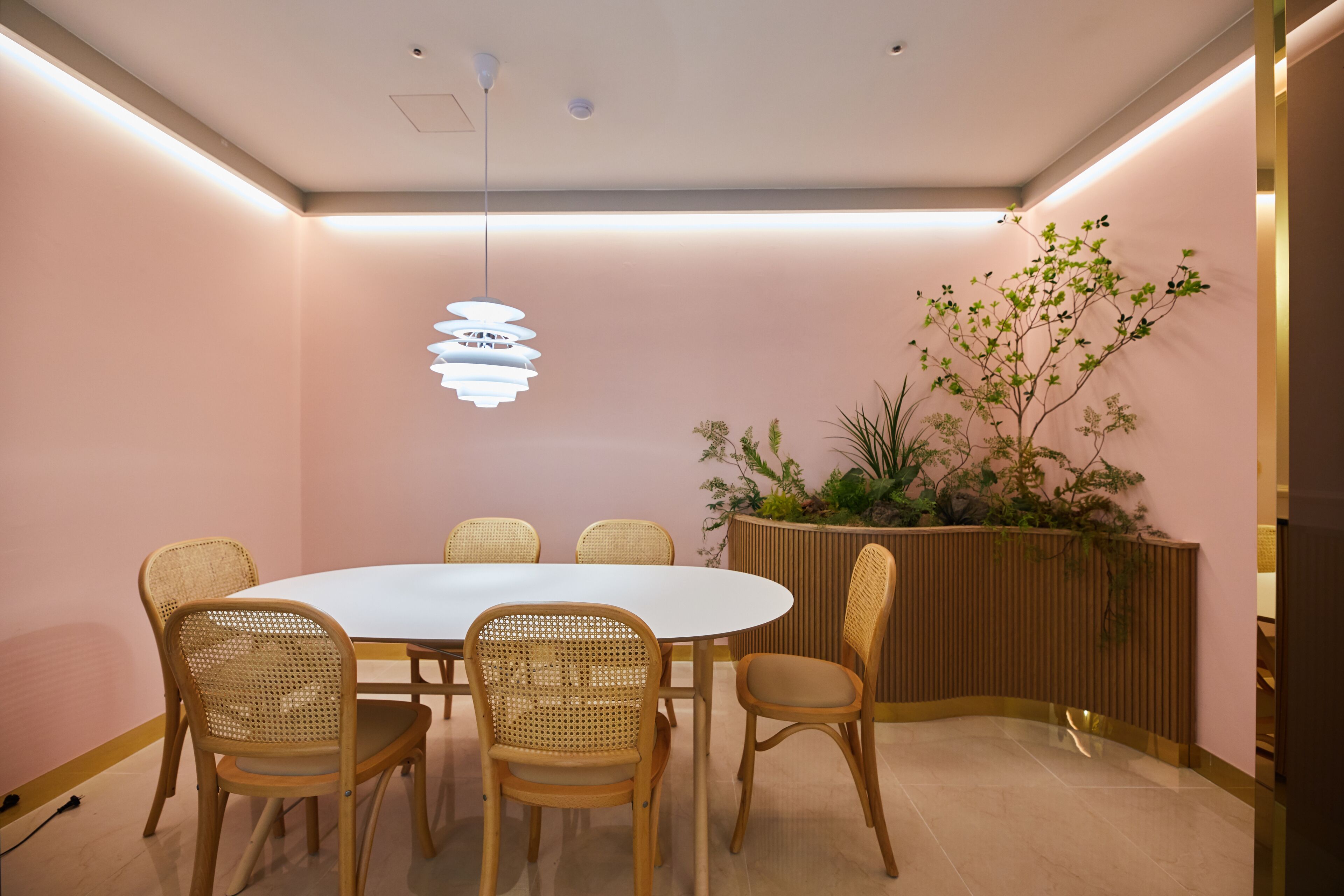 Uma sala de conferências com uma mesa redonda, seis cadeiras de vime, uma luminária pendente moderna e um vaso com planta.