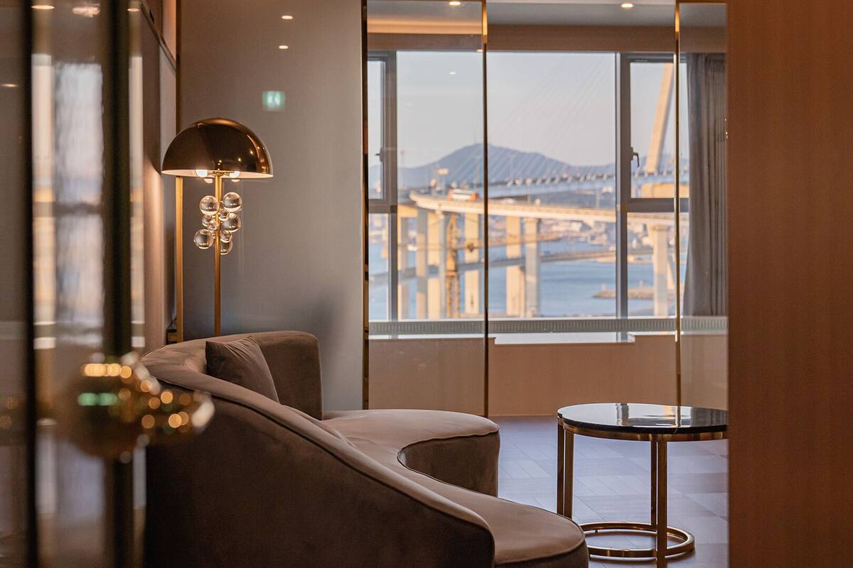 Un área de descanso moderna con sofá y una mesa pequeña, ventanas amplias que ofrecen vista a un puente y al paisaje urbano.