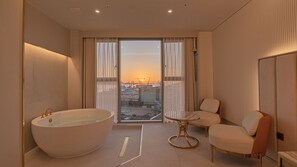 Un baño moderno con una bañera grande, una mesita redonda pequeña y una silla, con vistas a un paisaje urbano al atardecer.