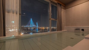 Una habitación de hotel moderna con un ventanal grande que ofrece una vista nocturna de un puente iluminado y luces de la ciudad.
