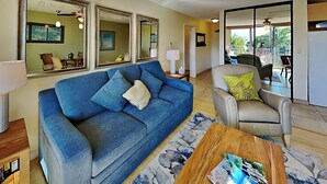 Una sala de estar con un sofá azul, un sillón gris y una mesa de centro de madera. Hay cuadros enmarcados en la pared y una ventana con vistas al exterior.