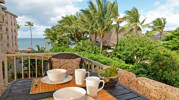 Una mesa de comedor dispuesta con vajilla y cubiertos blancos, con vistas a un paisaje tropical con palmeras y vista al océano.