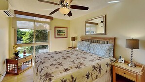 Un dormitorio con una cama grande, un ventilador de techo, una ventana que ofrece vistas a zonas verdes, una mesita de noche con una lámpara y un aparador con un teléfono.