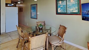 Un área de comedor con una mesa de vidrio rodeada de sillas de mimbre, una planta pequeña en maceta y cuadros en la pared.