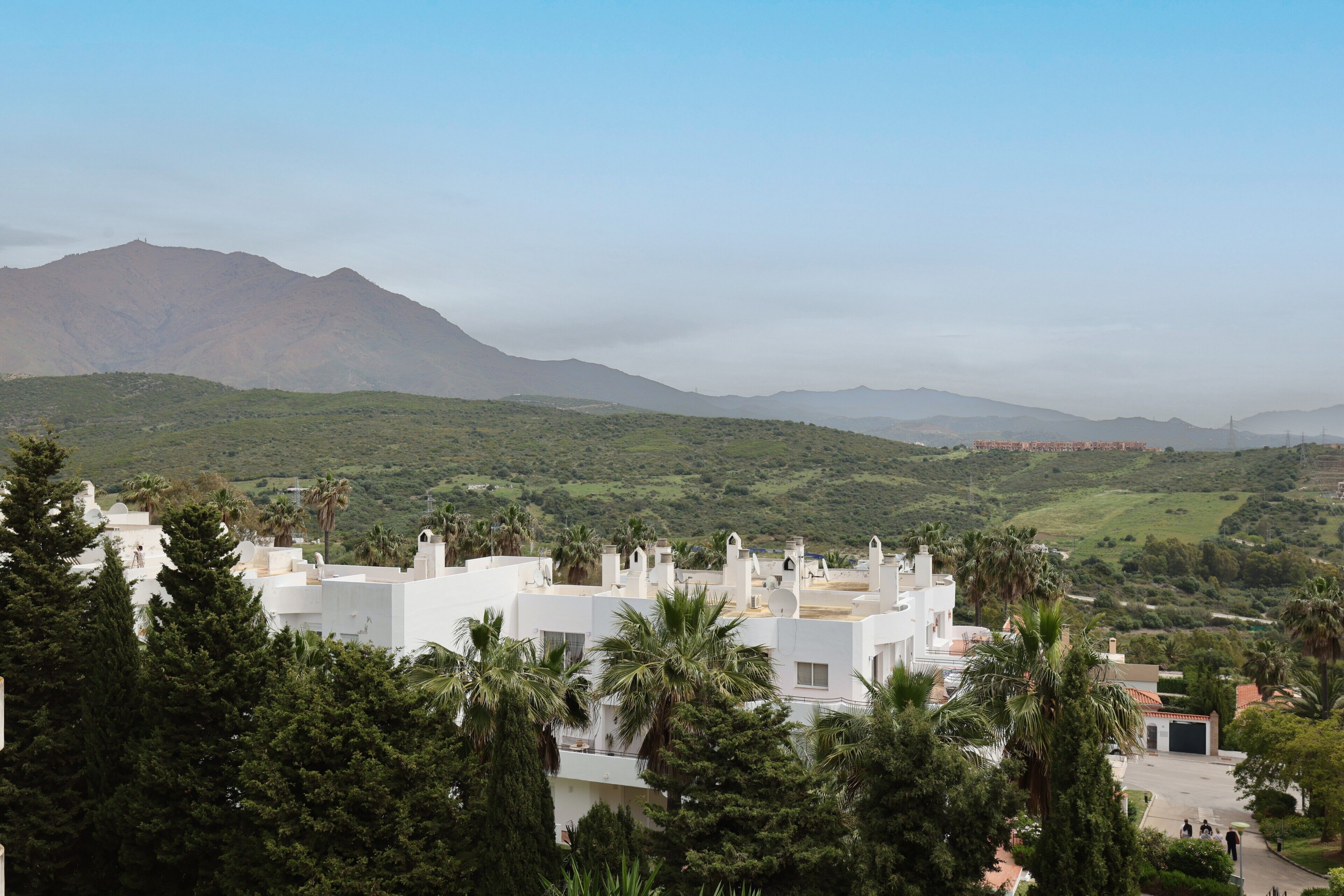 A white building with a flat roof, surrounded by greenery and palm trees, overlooking a hilly landscape.