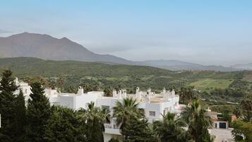A white building with a flat roof, surrounded by greenery and palm trees, overlooking a hilly landscape.
