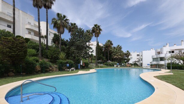 A swimming pool with a curved edge, surrounded by greenery and palm trees, with buildings in the background. - Apartamento "Del Paraíso" with Mountain View, Shared Pool and Private Terrace (Estepona)