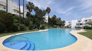 A swimming pool with a curved edge, surrounded by greenery and palm trees, with buildings in the background.