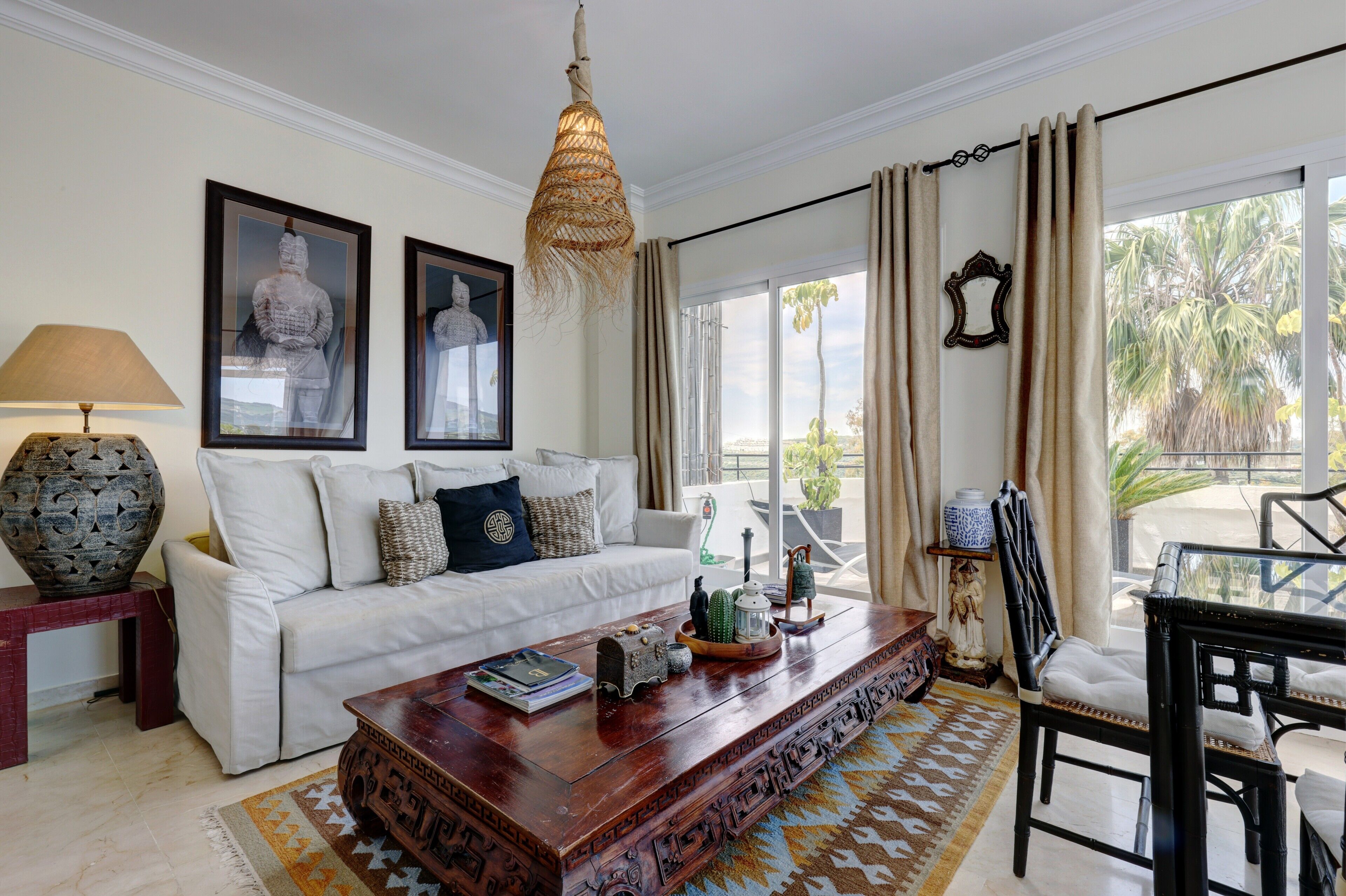 A living room with a white sofa, a wooden coffee table, a hanging light fixture, and a view of palm trees through the window.