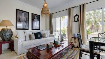 A living room with a white sofa, a wooden coffee table, a hanging light fixture, and a view of palm trees through the window.