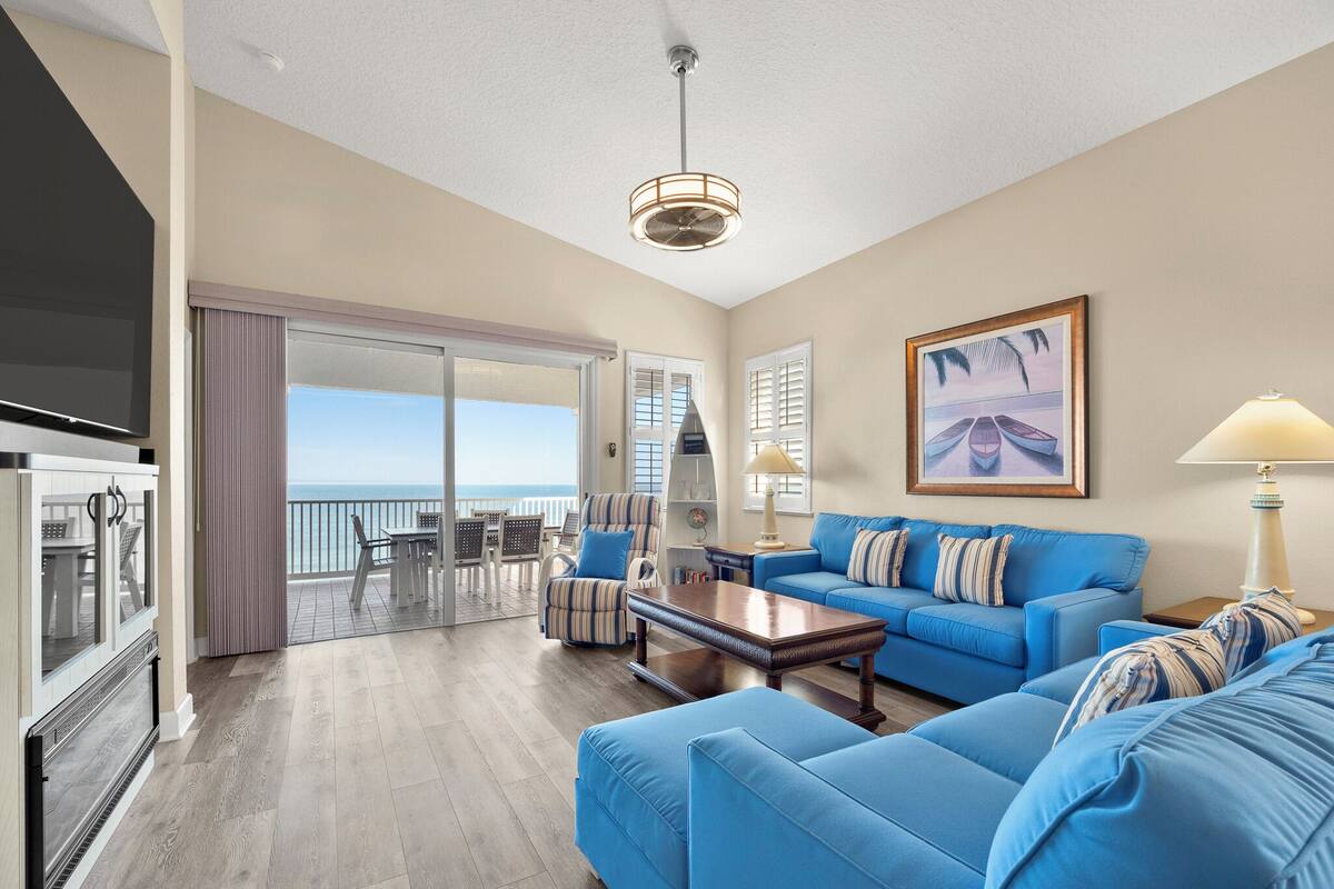 A spacious living area with a blue sofa set, a coffee table, and a TV. There is a balcony with a view of the ocean.