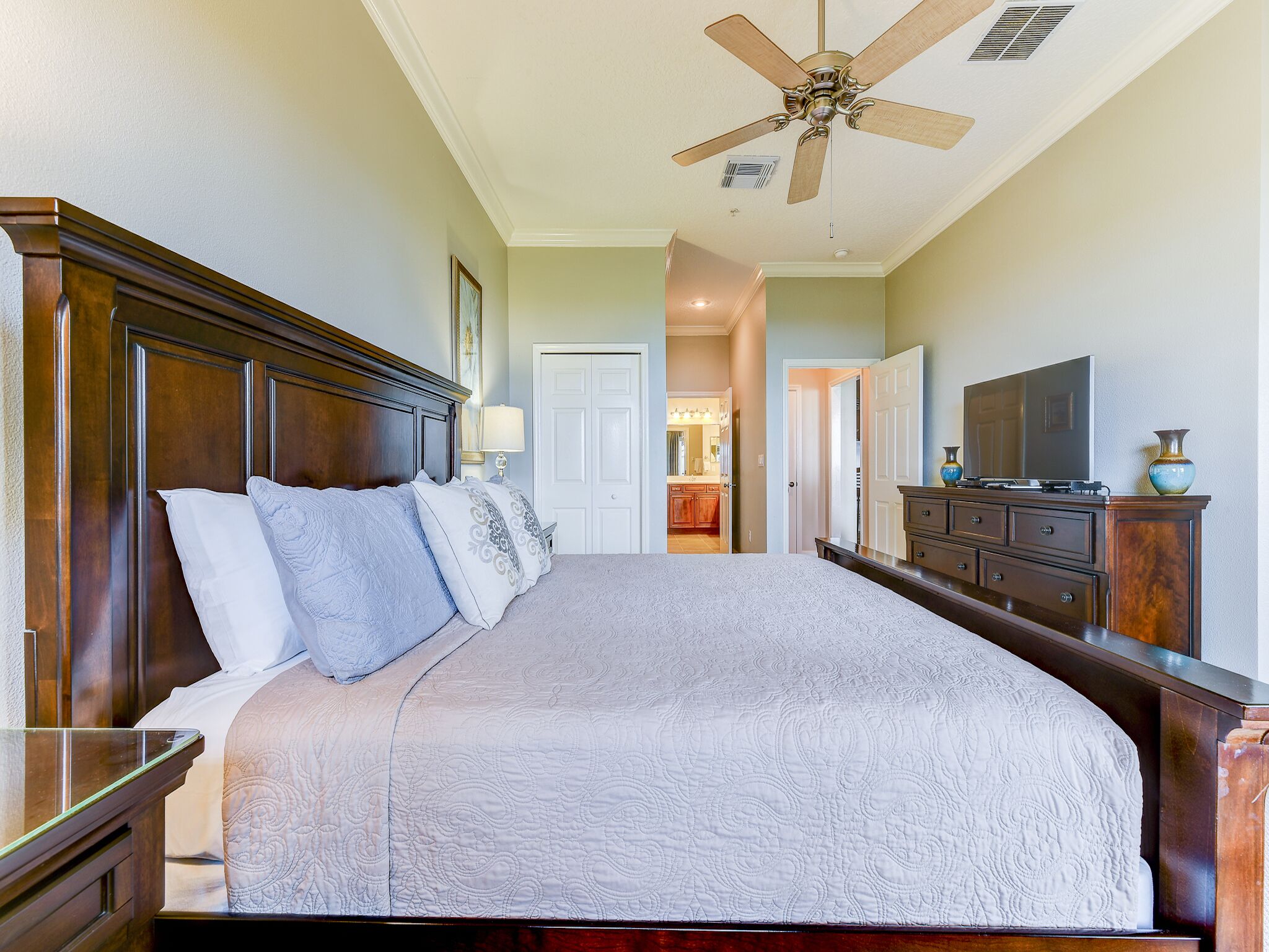 A bedroom with a large bed, wooden headboard, a ceiling fan, a dresser, and a television.