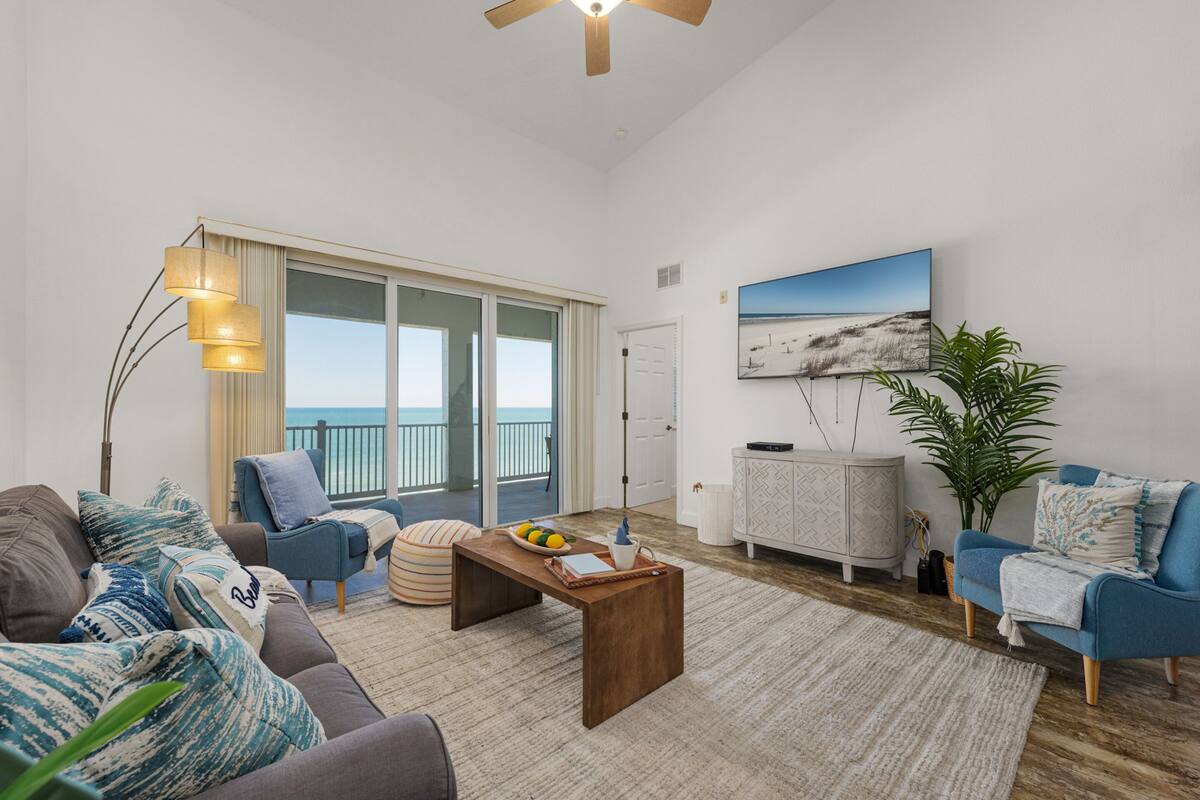 A spacious living room with a large window, a ceiling fan, a blue sofa, a wooden coffee table, and a potted plant.