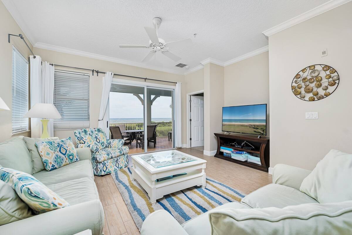A living room with a ceiling fan, a television, a sofa, a coffee table, and a view of the outdoors through large windows.