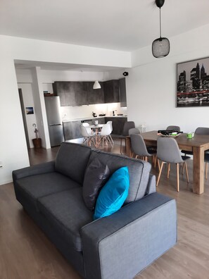 Ein modernes Wohnzimmer mit grauem Sofa, einem Esstisch mit Stühlen, einer Küchenzeile und einem Stadtbild an der Wand.