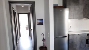 A modern kitchen with dark wooden flooring, a stainless steel refrigerator, and grey cabinets. A hallway leads to a bright room with a window. - Διαμέρισμα  (Αθηνα)