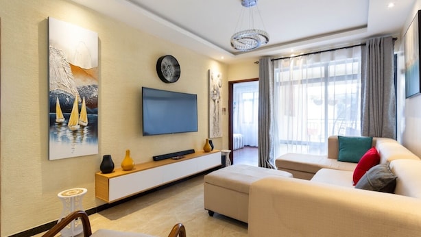 A modern living room with a flat-screen TV, a sofa, a coffee table, and a dining area with chairs. - Stay.Plus Dennis Garden Apartment (Nairobi)
