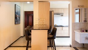 A modern kitchen with a central island, a dining area with a table and chairs, and a bathroom with a sink and cabinets. - Stay.Plus Dennis Garden Apartment (Nairobi)