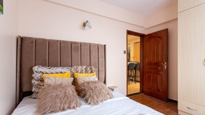 A bedroom with a large bed, a wooden door, and a small table. - Stay.Plus Dennis Garden Apartment (Nairobi)