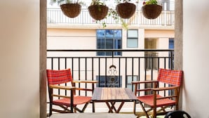 A balcony with a wooden table and two chairs, a metal railing, and hanging baskets with plants. - Stay.Plus Dennis Garden Apartment (Nairobi)