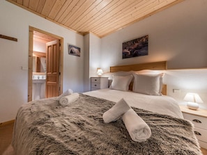 A bedroom with a wooden ceiling, a bed with a fluffy blanket, two pillows, a nightstand with a lamp, and a painting on the wall.