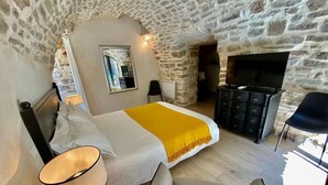 Une chambre avec un plafond en pierre, un lit recouvert d’un plaid jaune, une télévision, une commode et une chaise.
