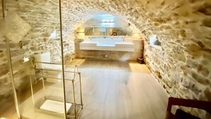 Une salle de bain aux murs en pierre, dotée d’une baignoire sur pieds, d’une douche à parois de verre et d’un lavabo blanc.
