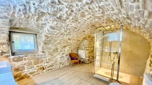 Une pièce rustique aux murs de pierre, dotée d’un plancher en bois, d’une fenêtre, d’un espace douche et de toilettes.