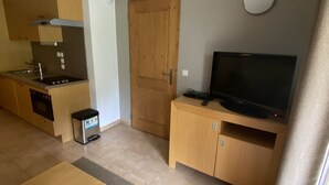 Un espai vital reduït amb petita cuina, una porta de fusta, un televisor sobre un suport de fusta i un petit cubell de brossa.