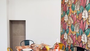 Una mesa de comedor lista con platos, cubiertos, vasos y una botella de vino, rodeada de sillas coloridas y una pared con un diseño de hojas vibrante.