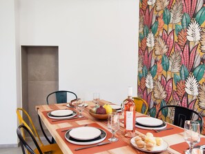 A dining table set with plates, cutlery, glasses, and a bottle of wine, surrounded by colorful chairs and a vibrant leaf-patterned wall.