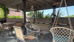 Un área de patio al aire libre con mesas y sillas de madera, una silla colgante y vistas a un edificio con tejado de tejas rojas.