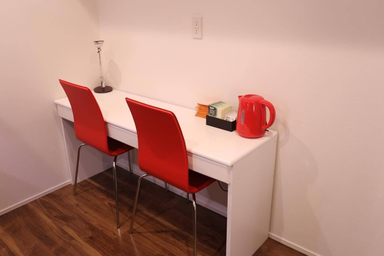 Uma pequena mesa branca com duas cadeiras vermelhas, uma cafeteira vermelha e uma luminária.
