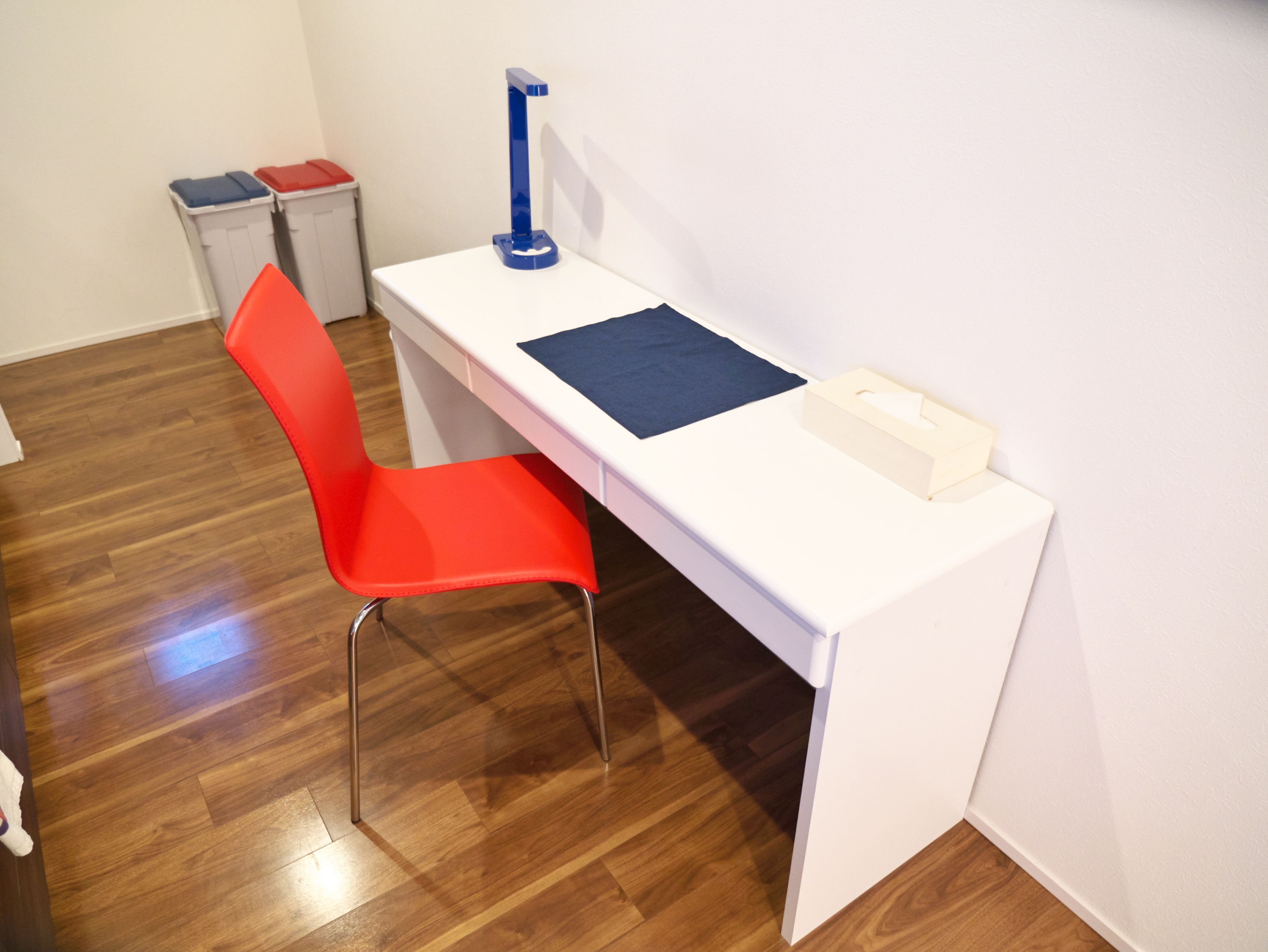 Een wit bureau met een blauwe lamp, een rode stoel en een kleine prullenbak in een kamer met houten vloeren.