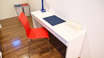 Een wit bureau met een blauwe lamp, een rode stoel en een kleine prullenbak in een kamer met houten vloeren.