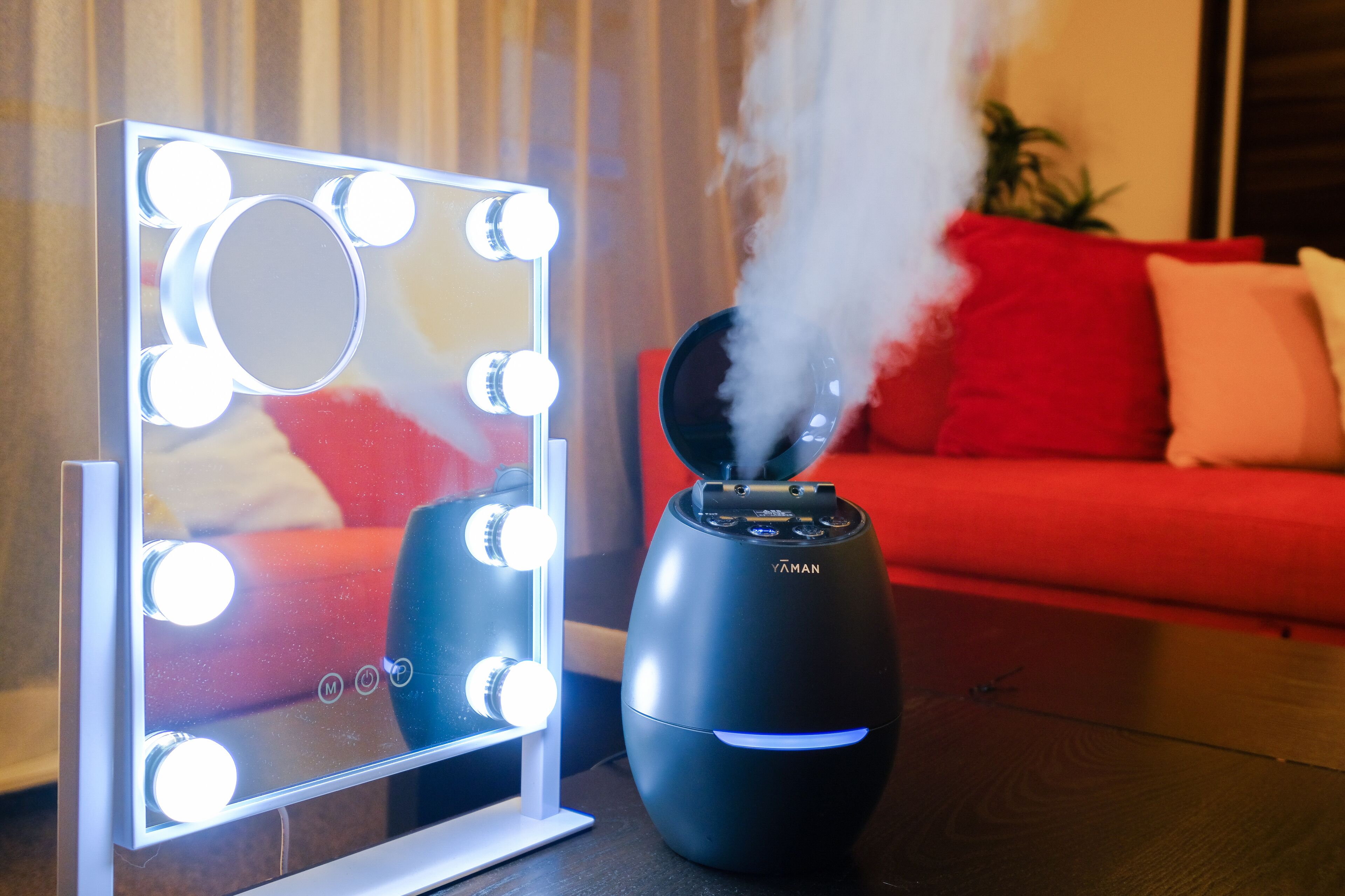 A mirror with lights, a black device with steam, and a red couch.